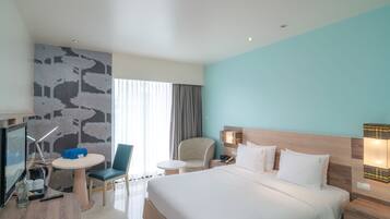 A hotel room with a large bed, a desk with a computer, a chair, a small table, and a TV.