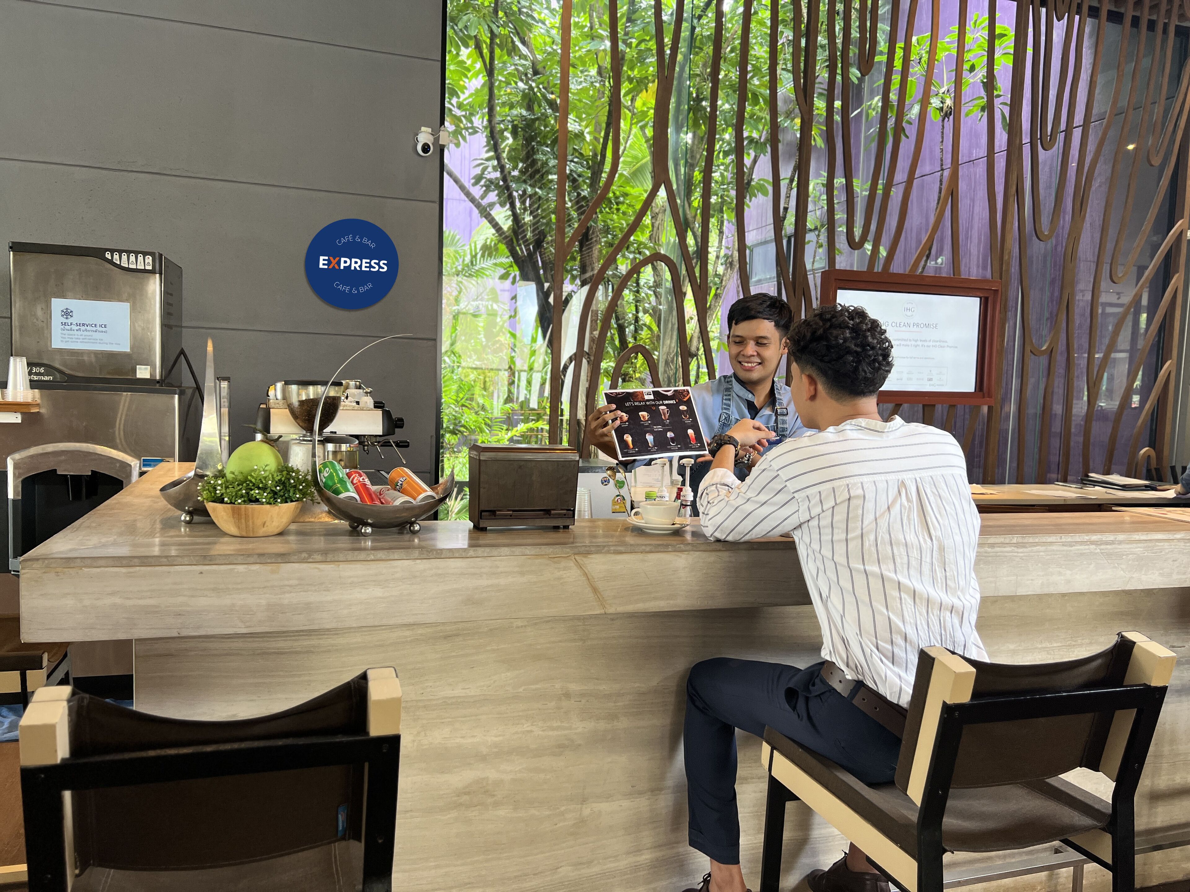 Een modern café met een zelfbedieningskiosk, een menubord en uitzicht op groen door het raam.