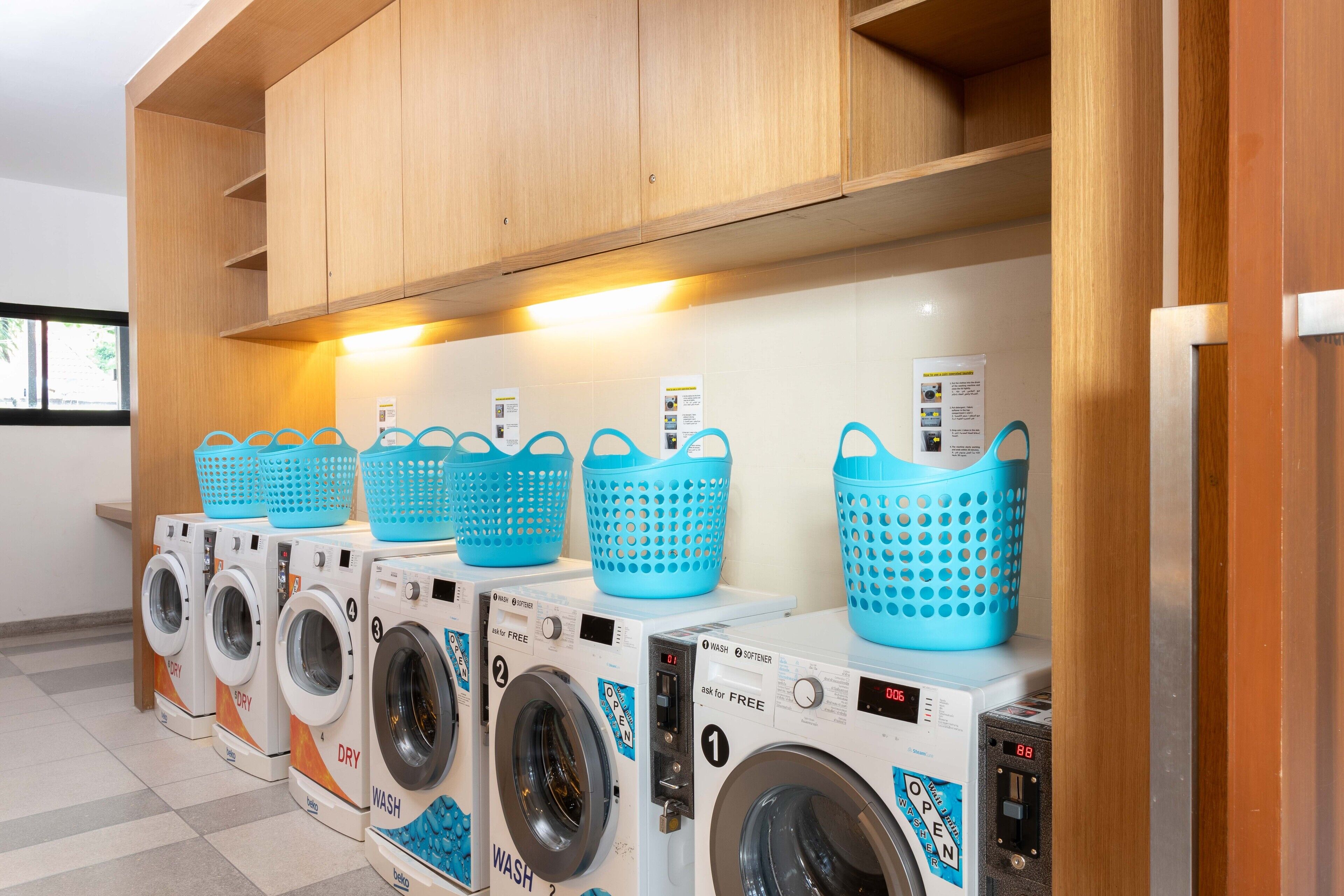 A row of front-loading washing machines with blue baskets on top, in a well-lit room with wooden cabinetry above.