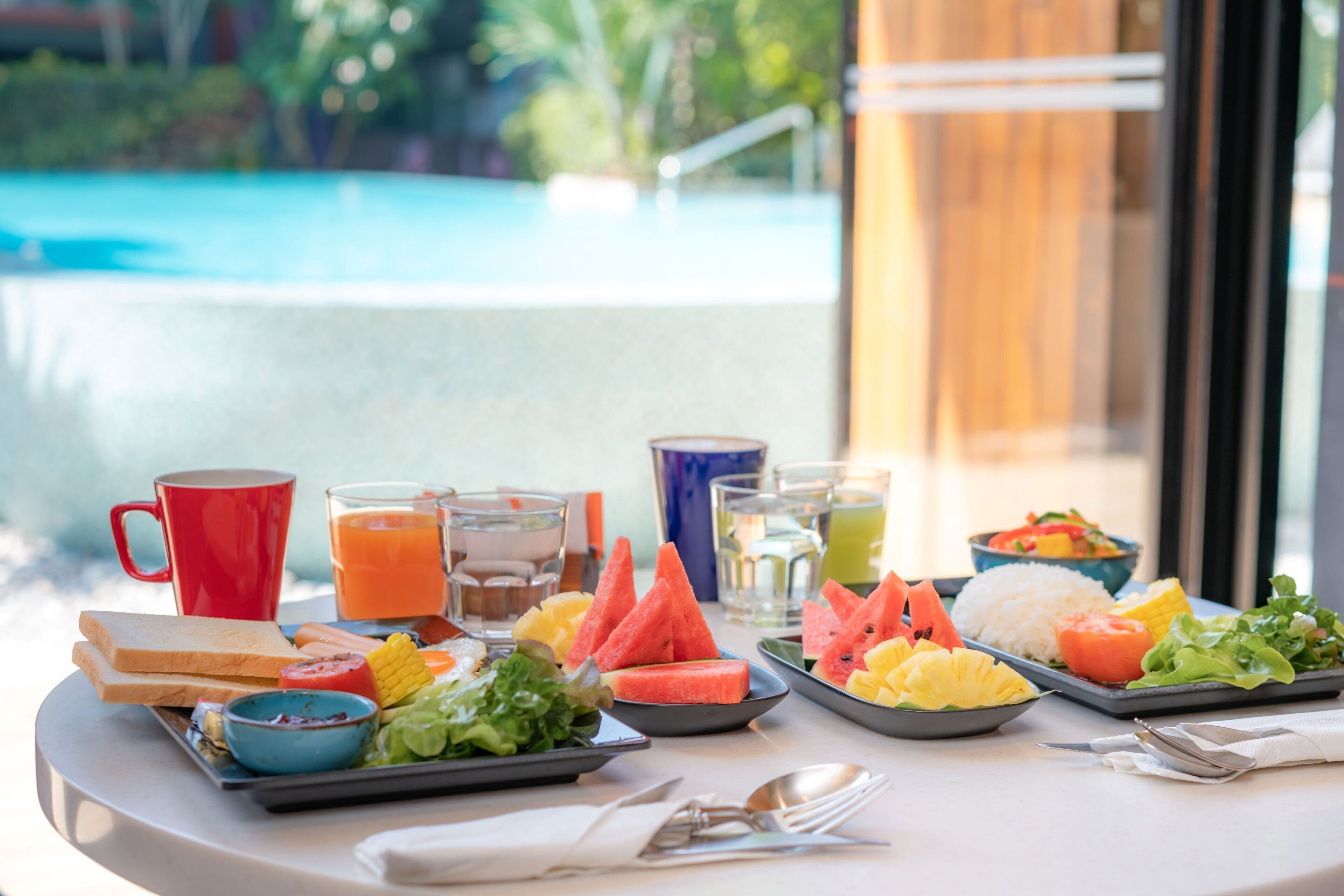 A breakfast buffet with various food items, drinks, and a red mug on a table with a pool view.