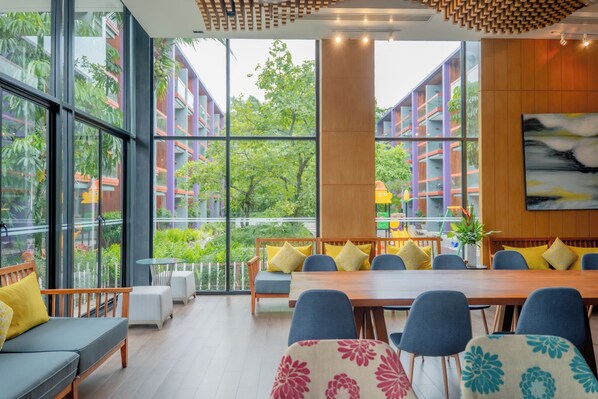 A modern dining area with a long wooden table, blue chairs, and yellow cushions. Large windows offer a view of greenery and buildings outside. - Holiday Inn Express Phuket Patong Beach Central by IHG (Patong)
