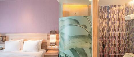 A hotel room with a bed, a nightstand, a lamp, a shower area, and a bathroom.