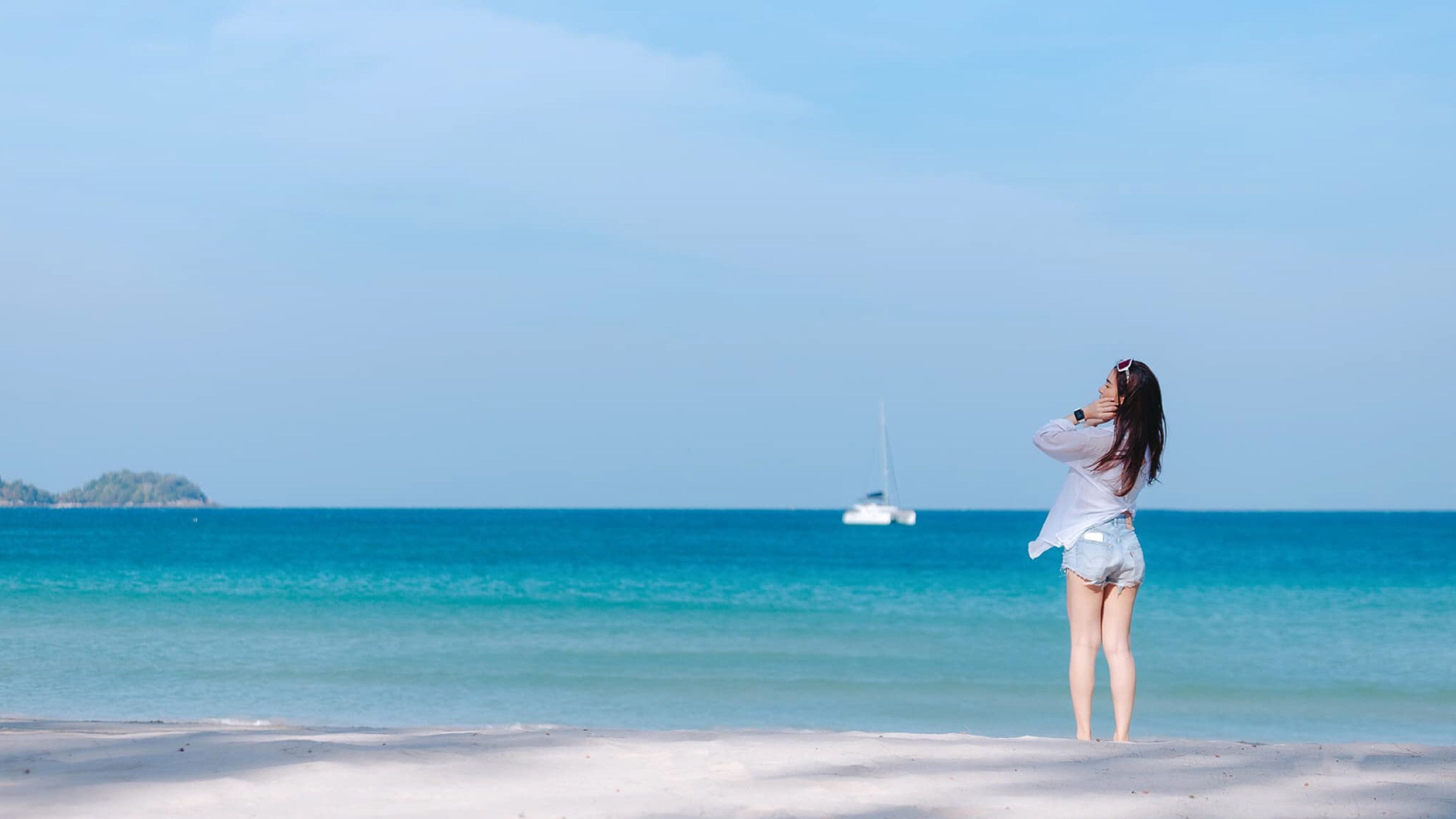 Eine Person steht am Strand mit freiem Blick auf das Meer und einem Segelboot in der Ferne.