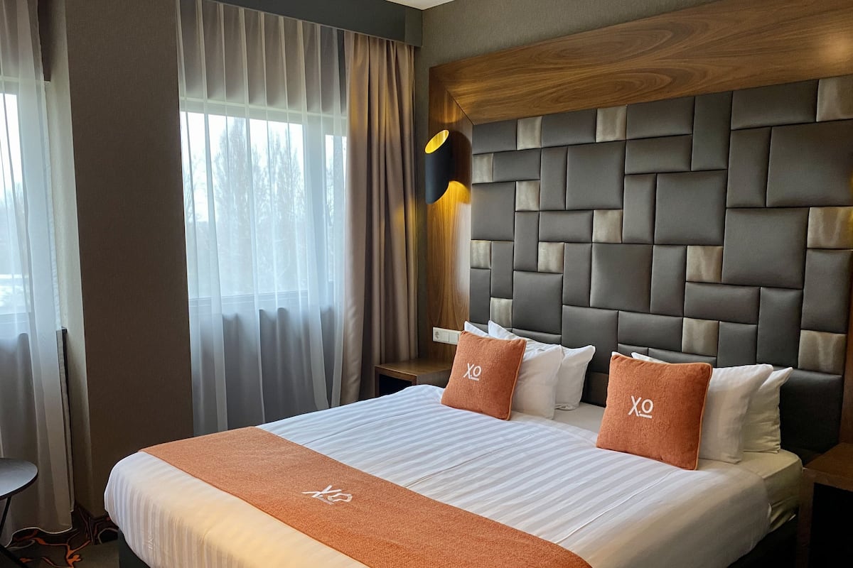 Une chambre d’hôtel avec un grand lit, une tête de lit ornée d’un motif géométrique et une table de chevet avec une lampe.