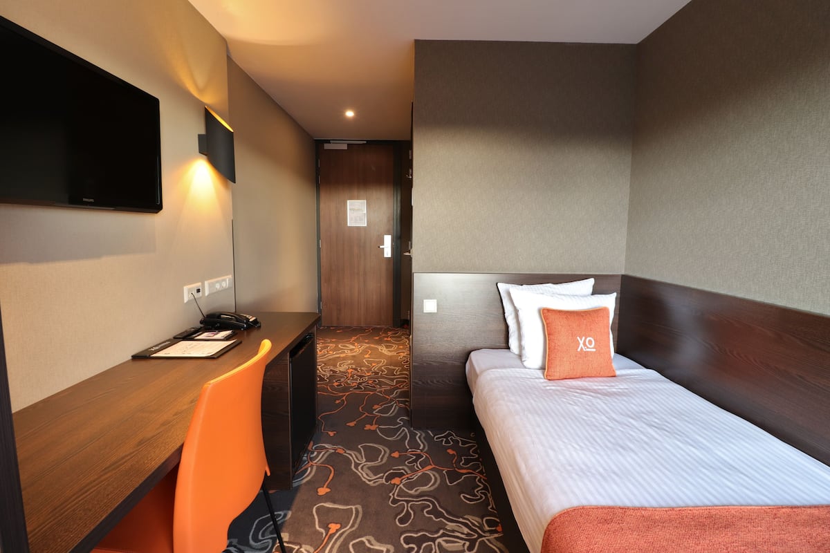 Une chambre d’hôtel avec un lit, un bureau, une chaise orange, un téléviseur à écran plat fixé au mur et un sol recouvert de moquette.