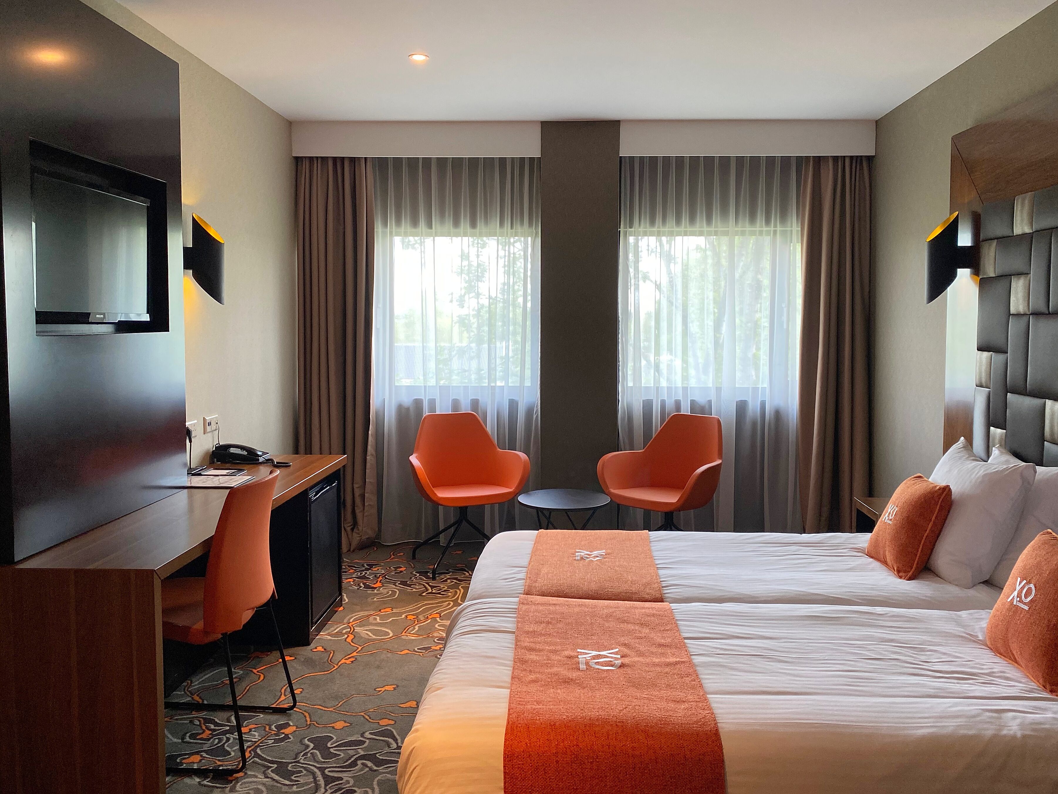 A hotel room with a bed, two orange chairs, a small table, a desk, a TV, and a large window with curtains.