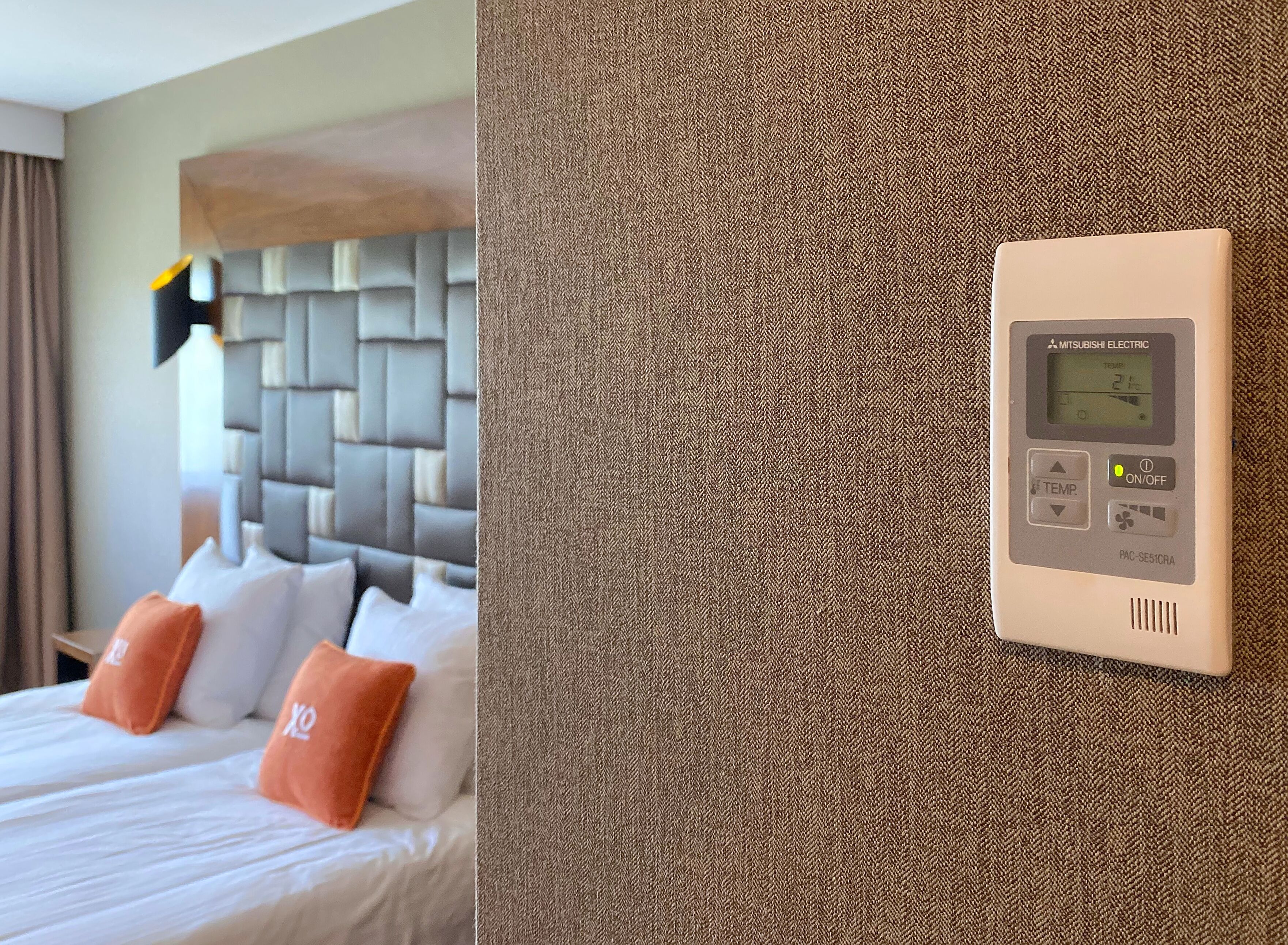 A hotel room with a bed, pillows, and a wall-mounted thermostat.