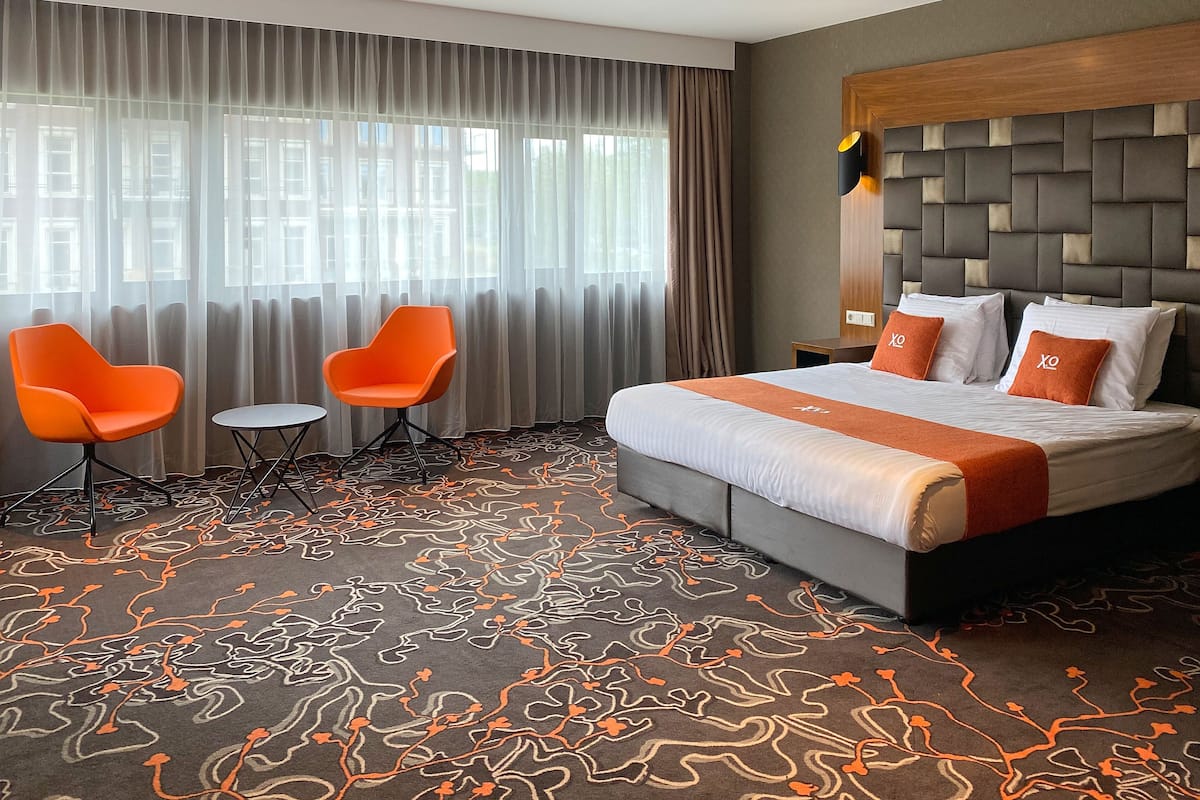 Une chambre d’hôtel avec un grand lit, deux fauteuils orange, une petite table et un tapis à motifs.