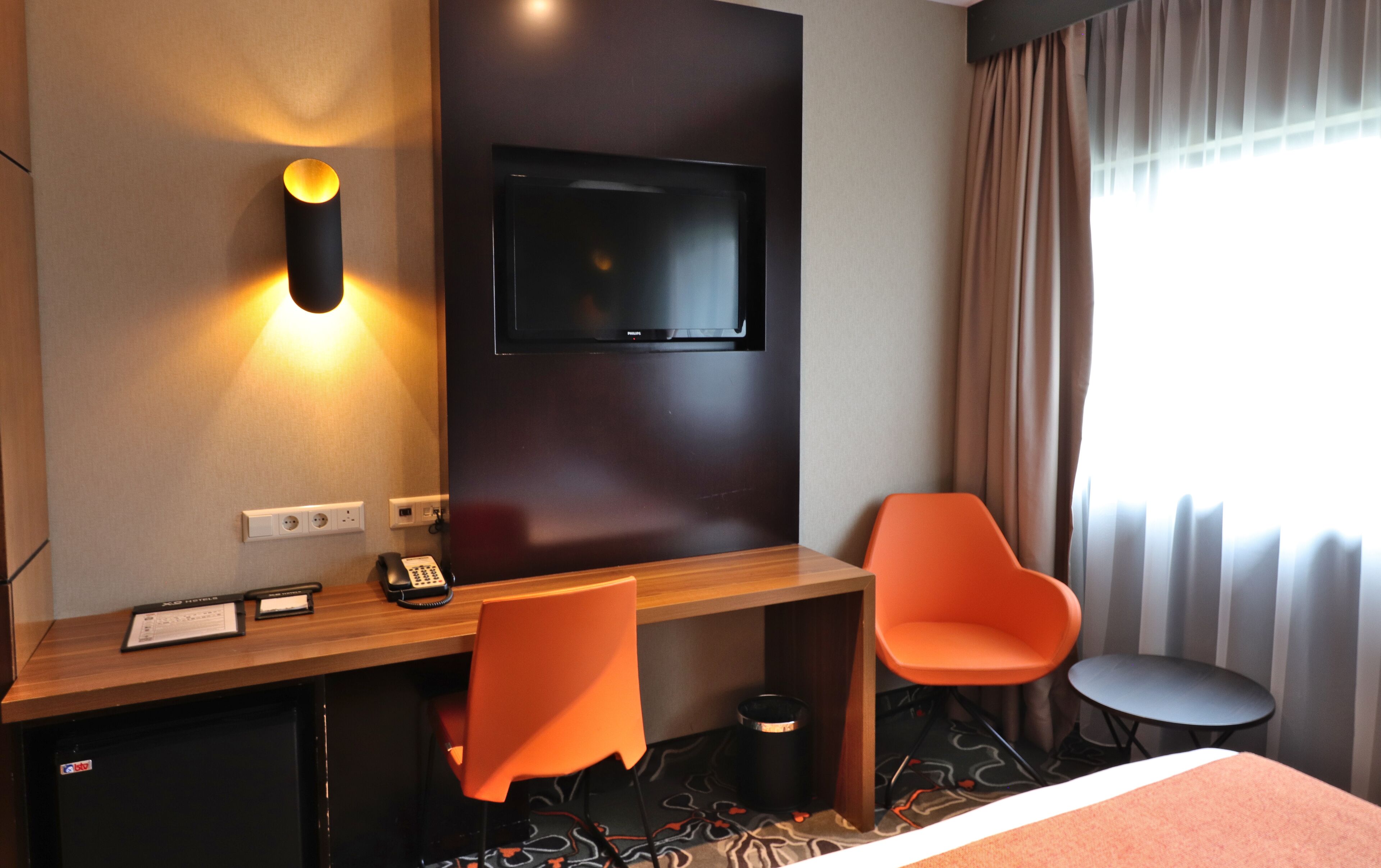 Habitación de hotel con escritorio de madera, dos sillas color naranja, televisor montado en la pared, cama, mesita y ventana con cortinas.