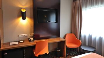 Habitación de hotel con escritorio de madera, dos sillas color naranja, televisor montado en la pared, cama, mesita y ventana con cortinas.