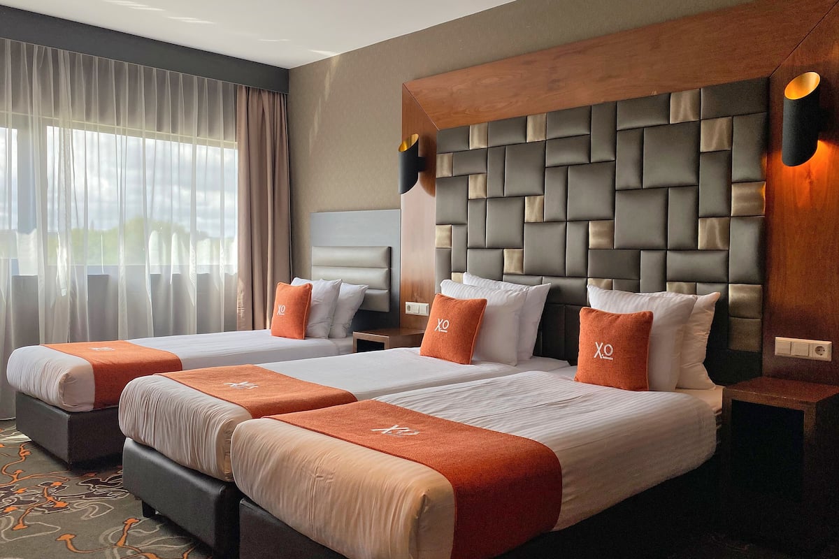 Une chambre d’hôtel avec deux lits, une grande tête de lit rembourrée et une fenêtre ornée de rideaux légers.