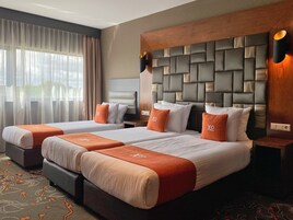 A hotel room with two beds, a large padded headboard, and a window with sheer curtains.