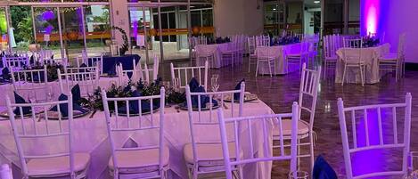Un salón de banquetes con mesas redondas dispuestas para comer, sillas blancas y luces colgantes en el techo.
