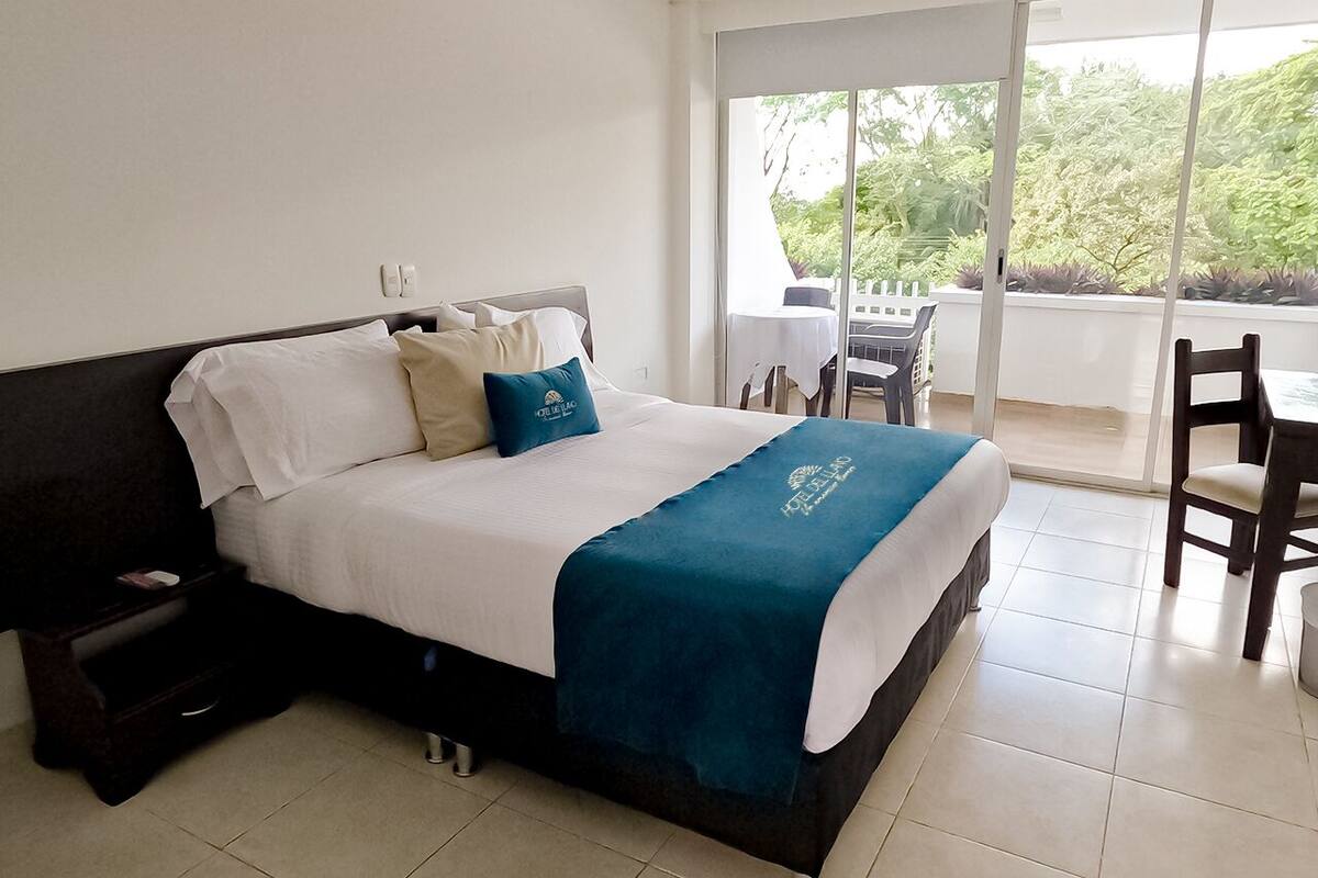 Une chambre d’hôtel avec un lit, une table de chevet, une table avec des chaises et une vue sur la verdure par la fenêtre.