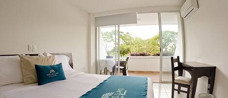 A hotel room with a bed, a desk, and a chair. There is a window with a view of greenery outside.