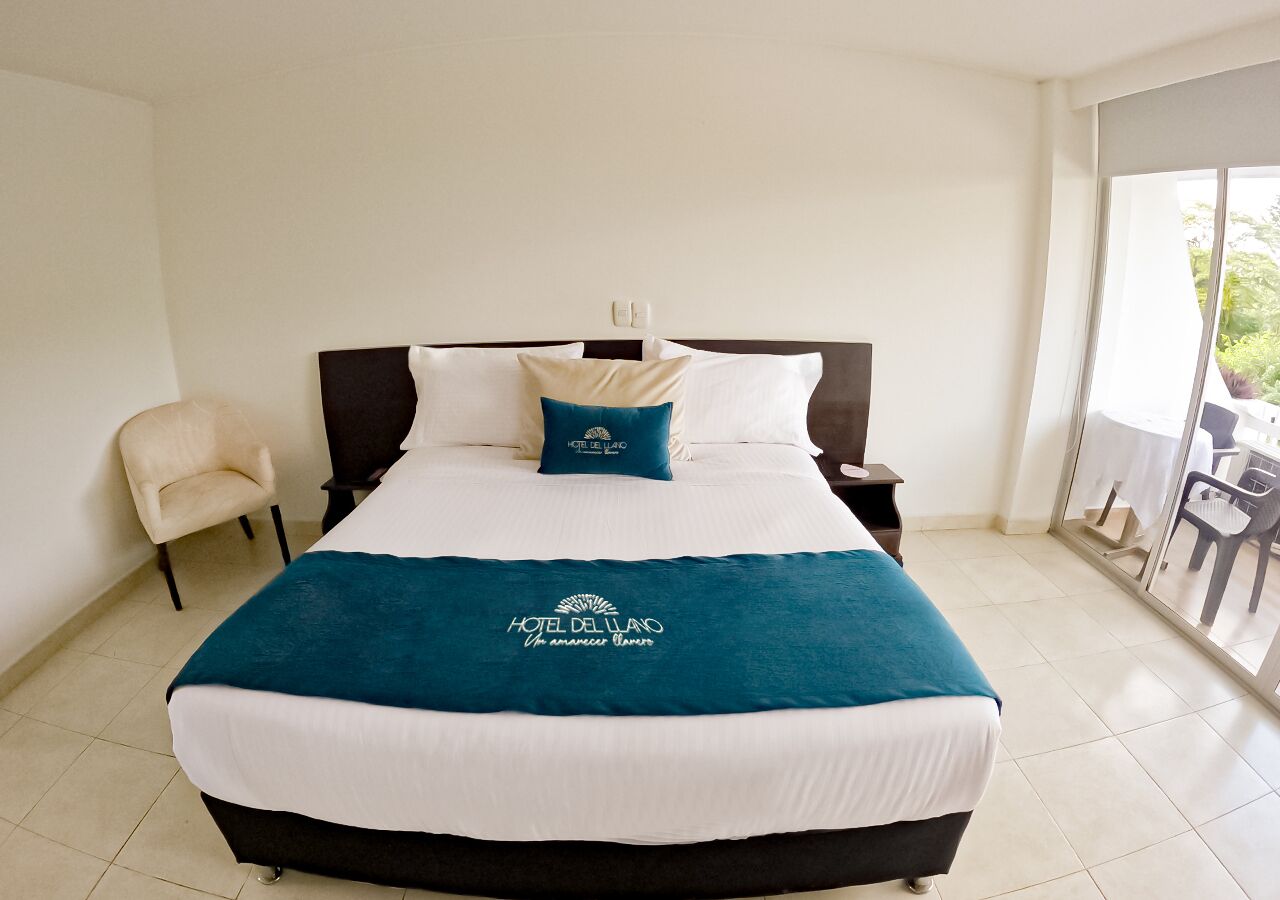 Habitación de hotel con una cama grande, una silla, una mesita de noche y vistas al exterior a través de un ventanal.