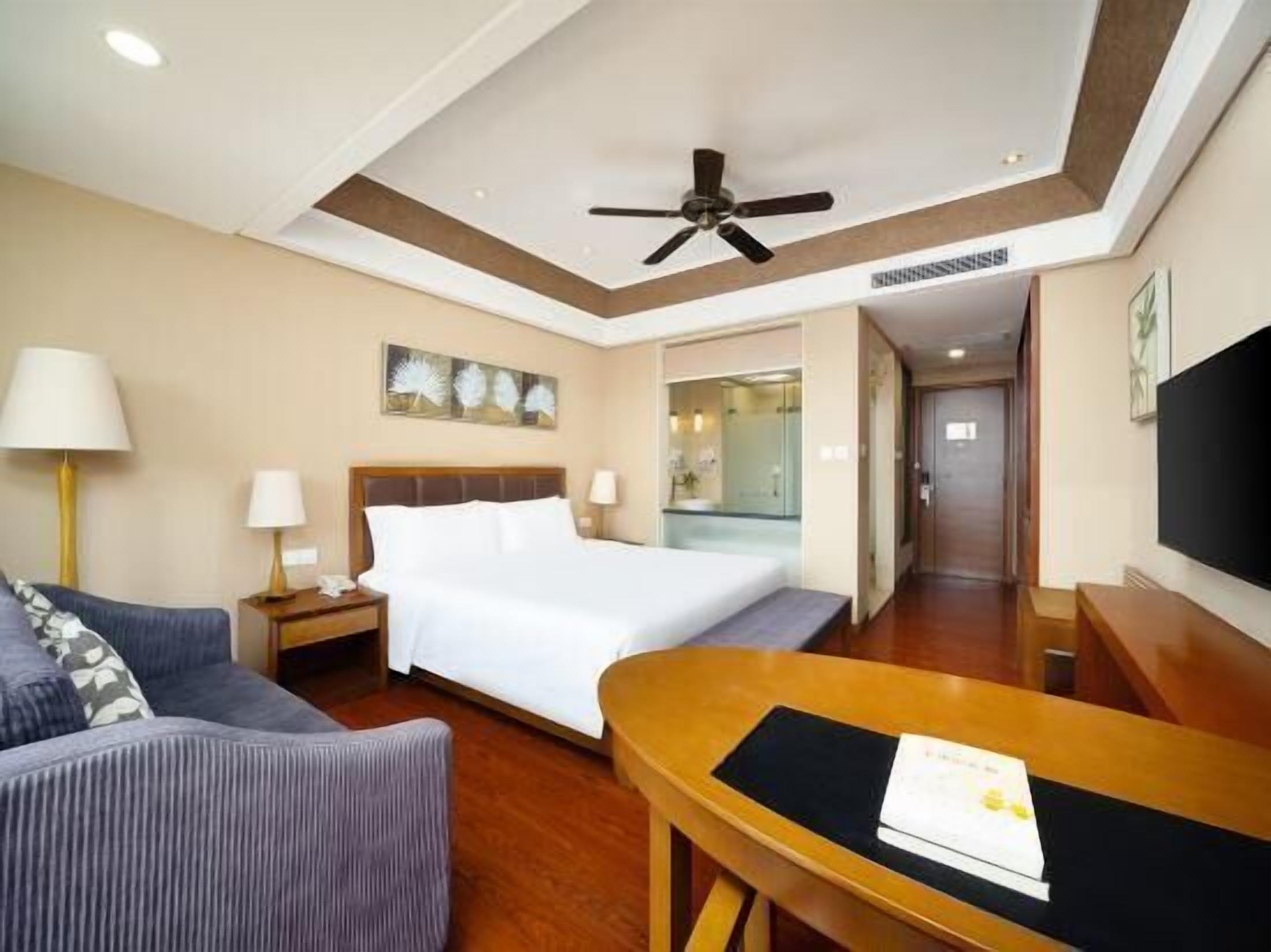 A hotel room with a bed, a desk, a chair, a sofa, a ceiling fan, and a TV.
