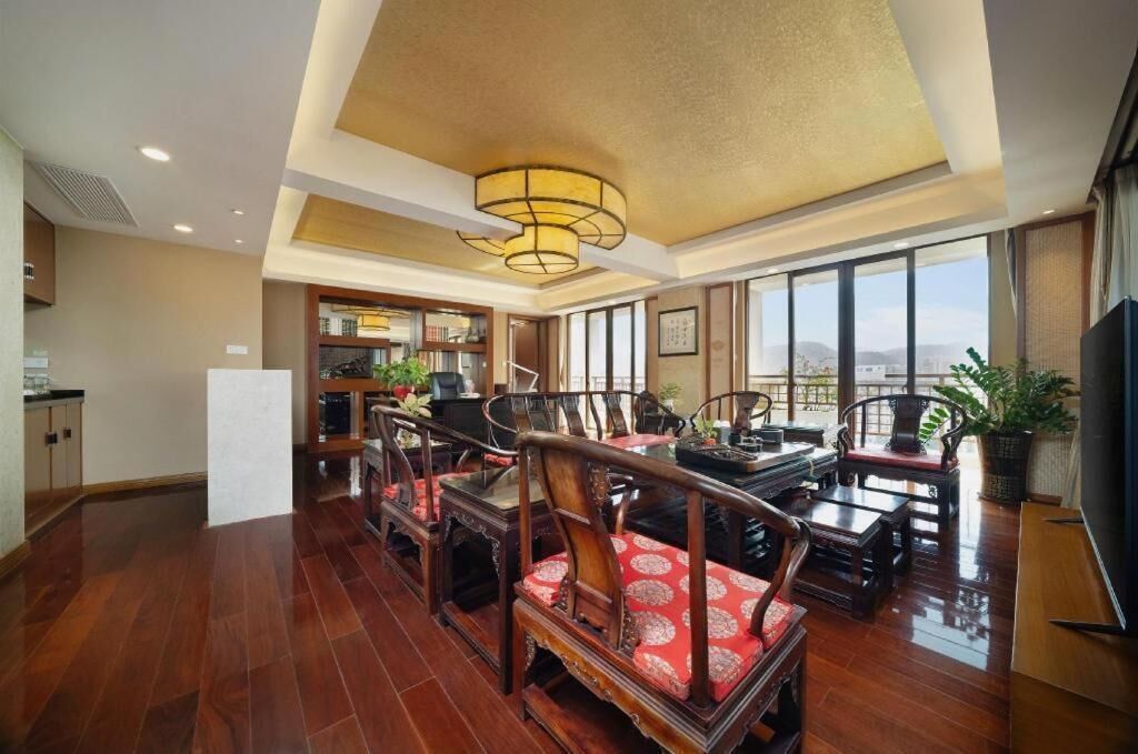 A spacious living area with a high ceiling, wooden furniture, and a large window offering a city view.