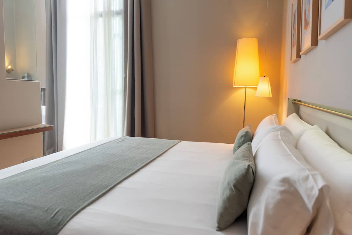 A neatly made bed with white linens and a grey throw blanket, a bedside lamp, and a view of a window with curtains.