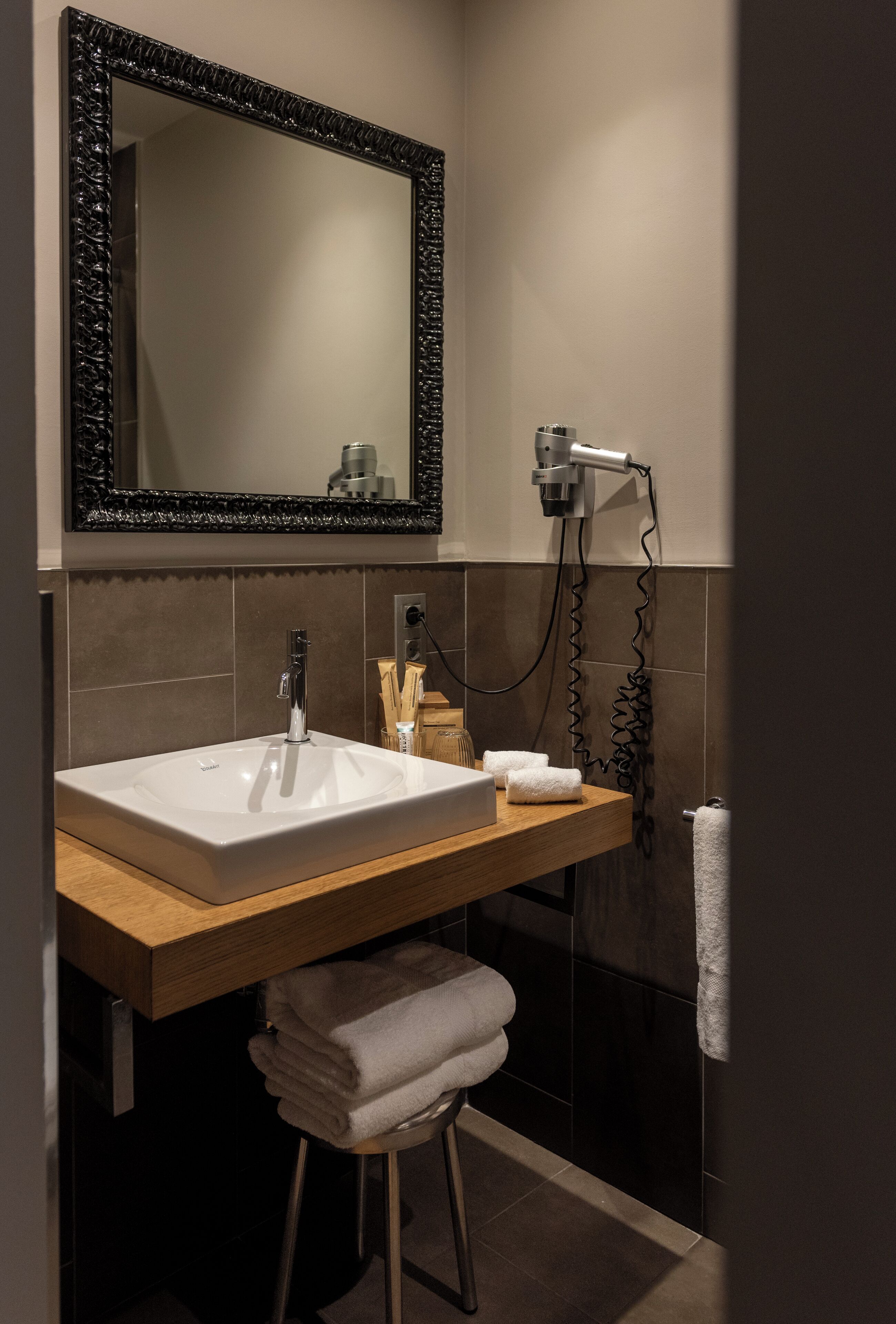 Un baño moderno con un lavamanos blanco, tocador de madera, un espejo grande y un secador de pelo en la pared.