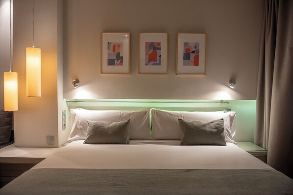 Habitació d'hotel amb un llit, almoines, una taula de nit, una làmpada i tres quadres al mur.
