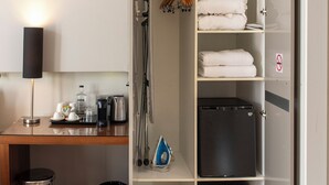 A hotel room with a desk, a lamp, a mini fridge, a trash bin, and a wardrobe with shelves. - Vasanta Hotel (Barcelona)