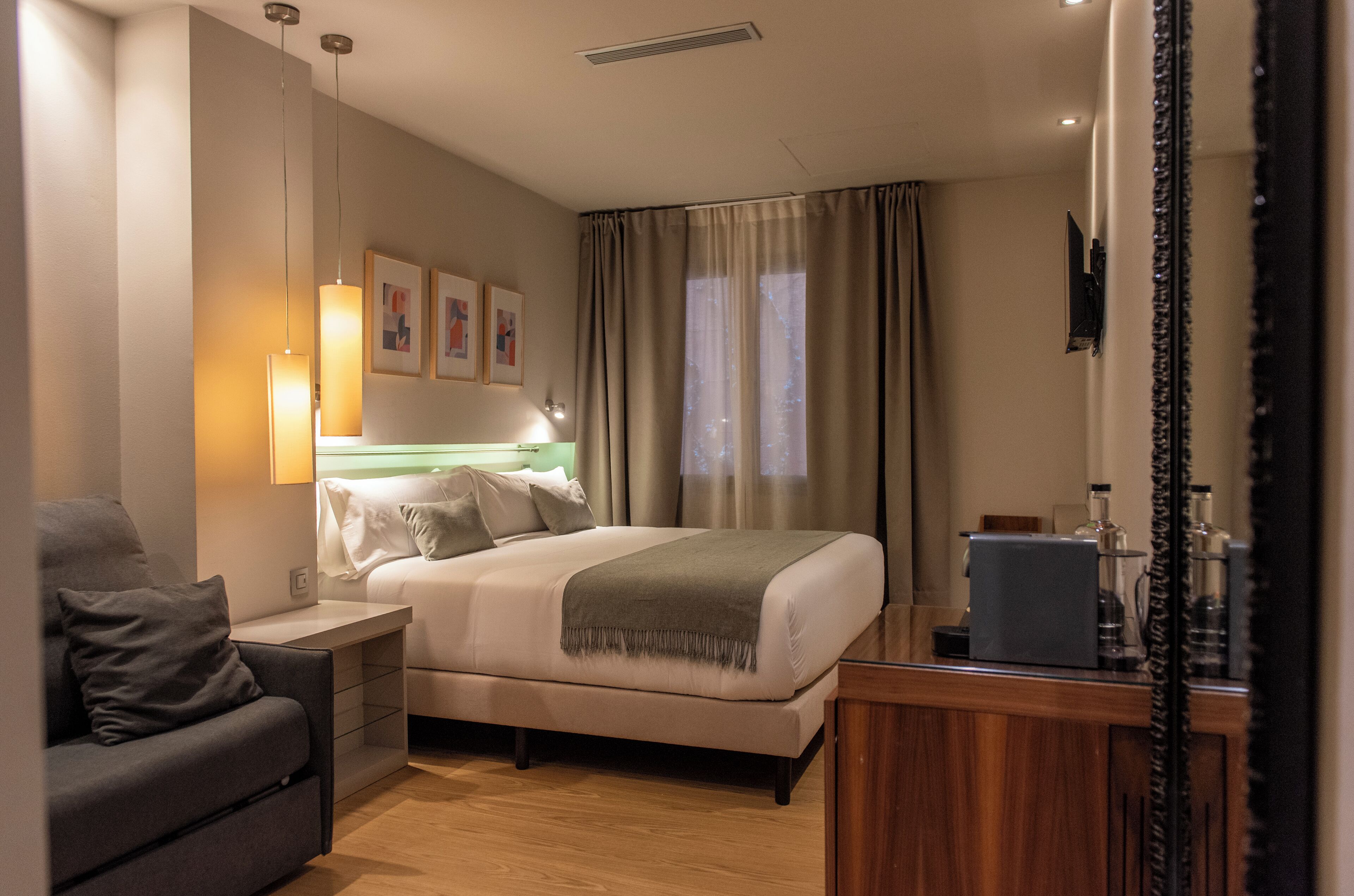 Habitación de hotel con cama, escritorio, silla, mesita de noche, lámpara, espejo y ventana con cortinas.