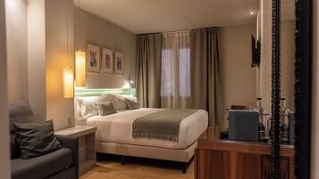 Habitación de hotel con cama, escritorio, silla, mesita de noche, lámpara, espejo y ventana con cortinas.
