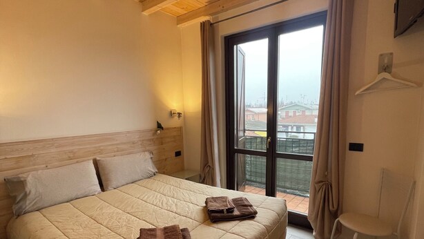 A bedroom with a bed, a chair, a window with curtains, and a wooden ceiling. - Homely - Malpensa & Fiere Milano (Cardano al Campo)