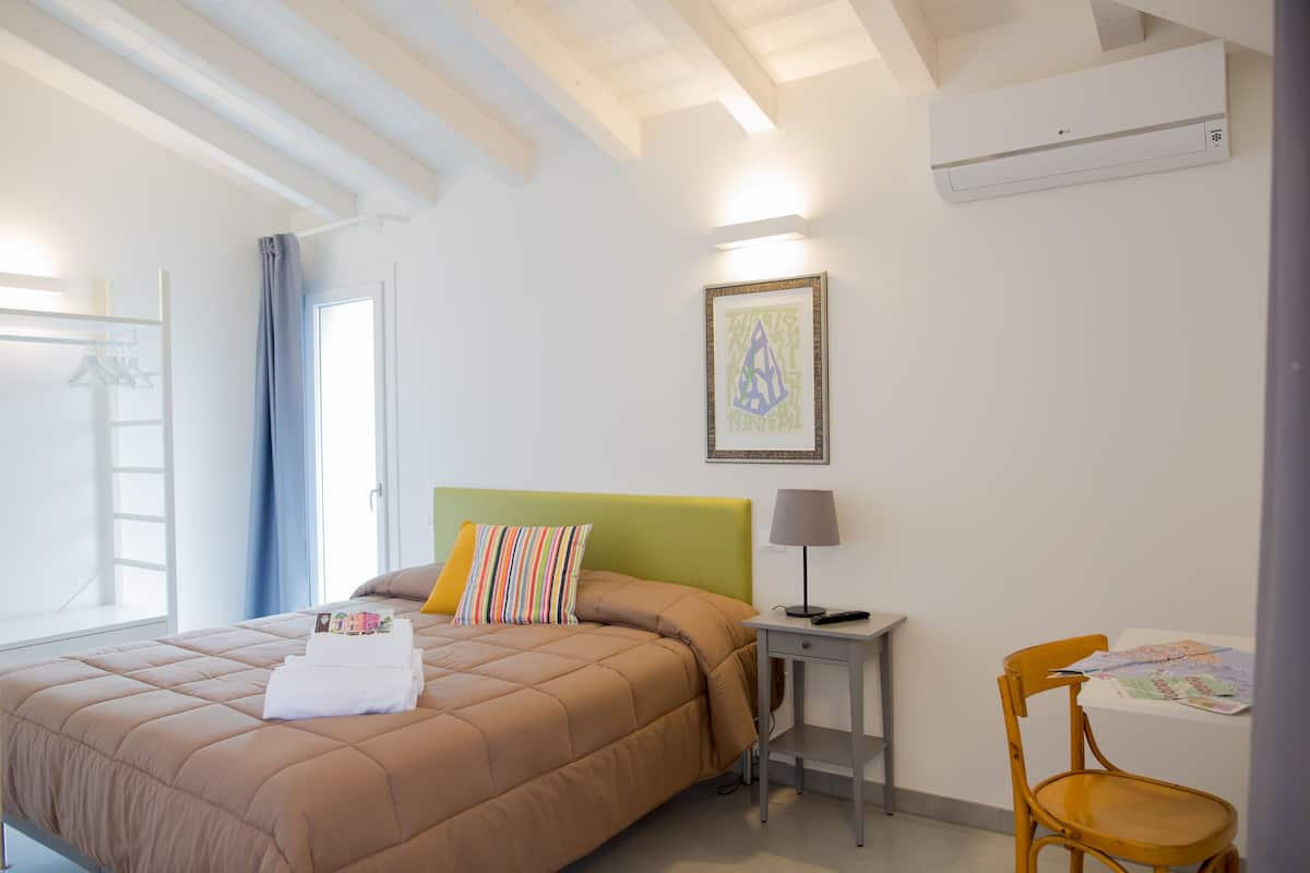 A neatly made bed with a brown comforter, a small bedside table with a lamp, a chair, and a framed picture on the wall.