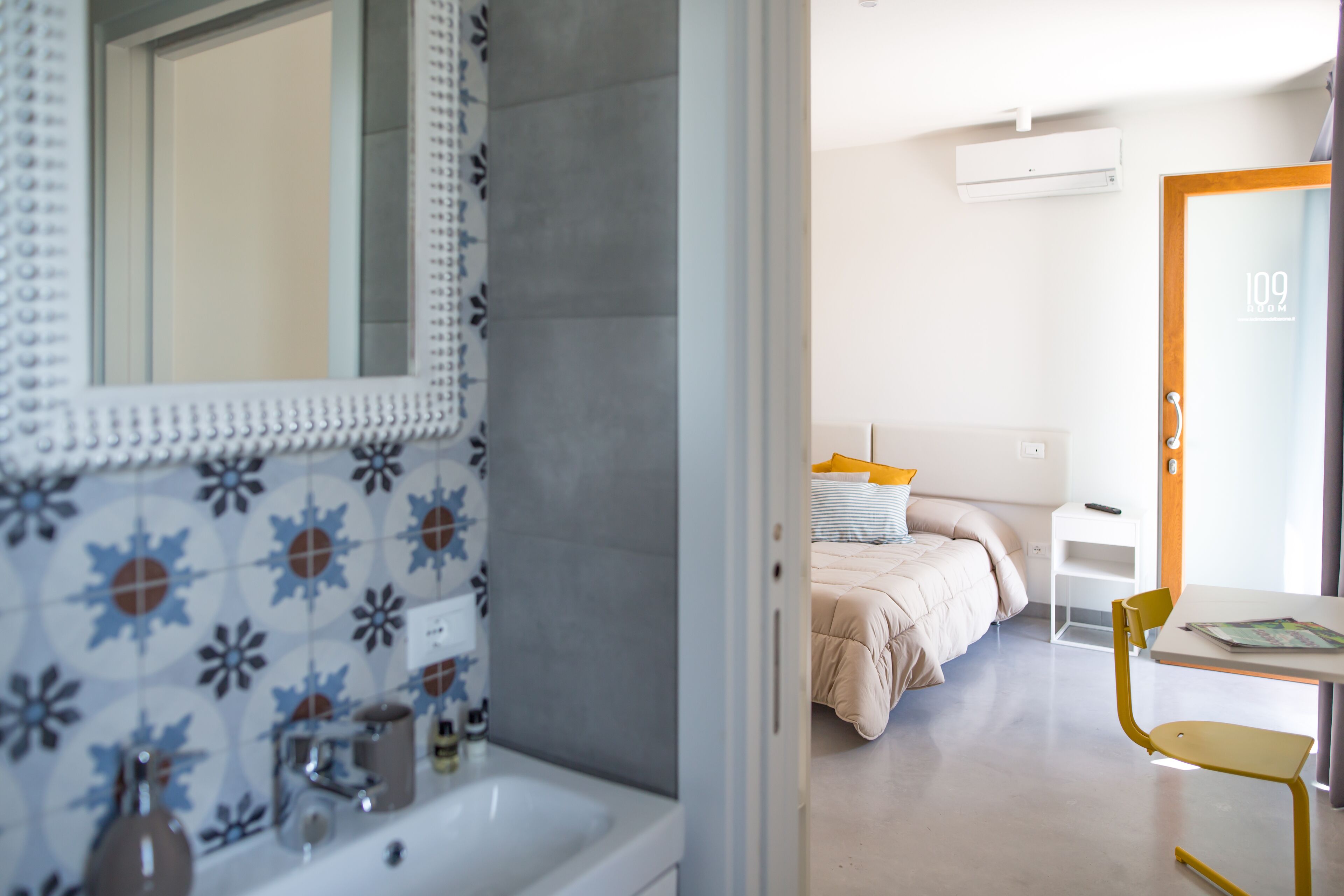 A modern hotel room with a bathroom, a bed, a small table, and a chair.