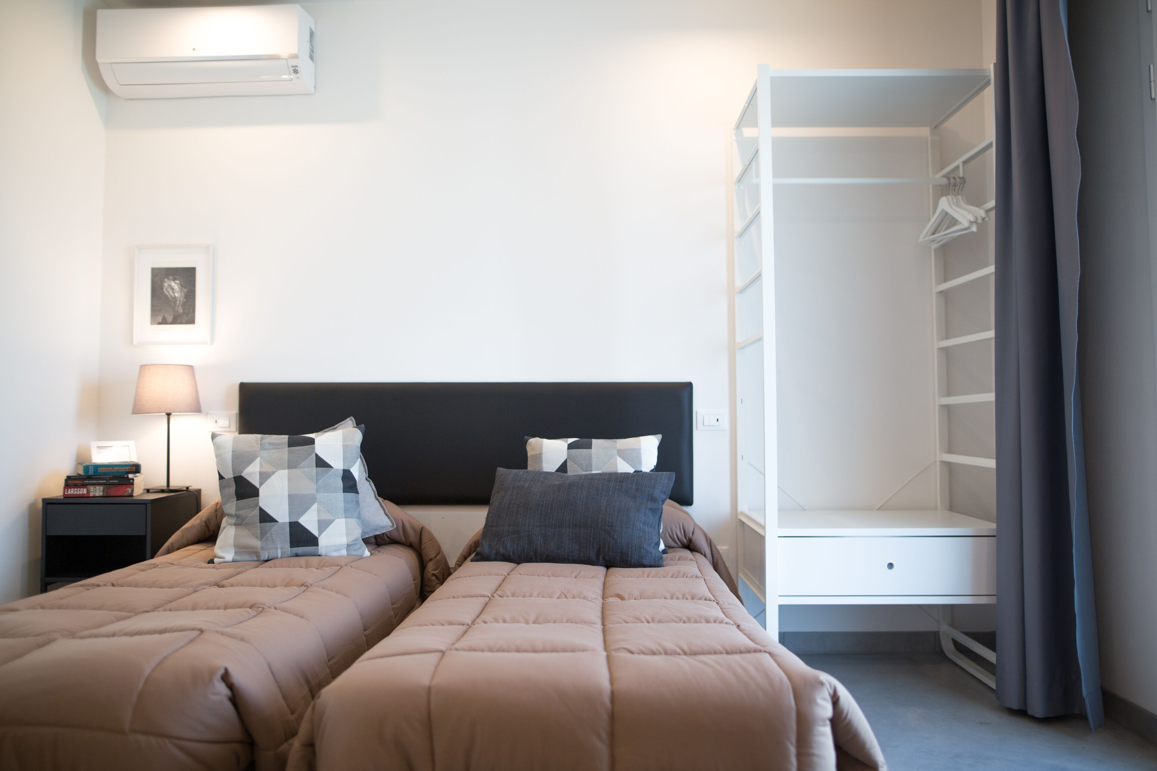 A hotel room with two beds, a bedside table with a lamp, a wardrobe, and a framed picture on the wall.