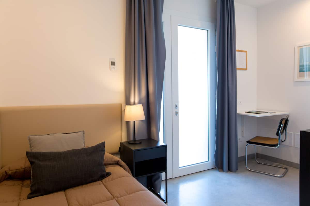 A hotel room with a bed, a desk, a chair, a lamp, and a door.