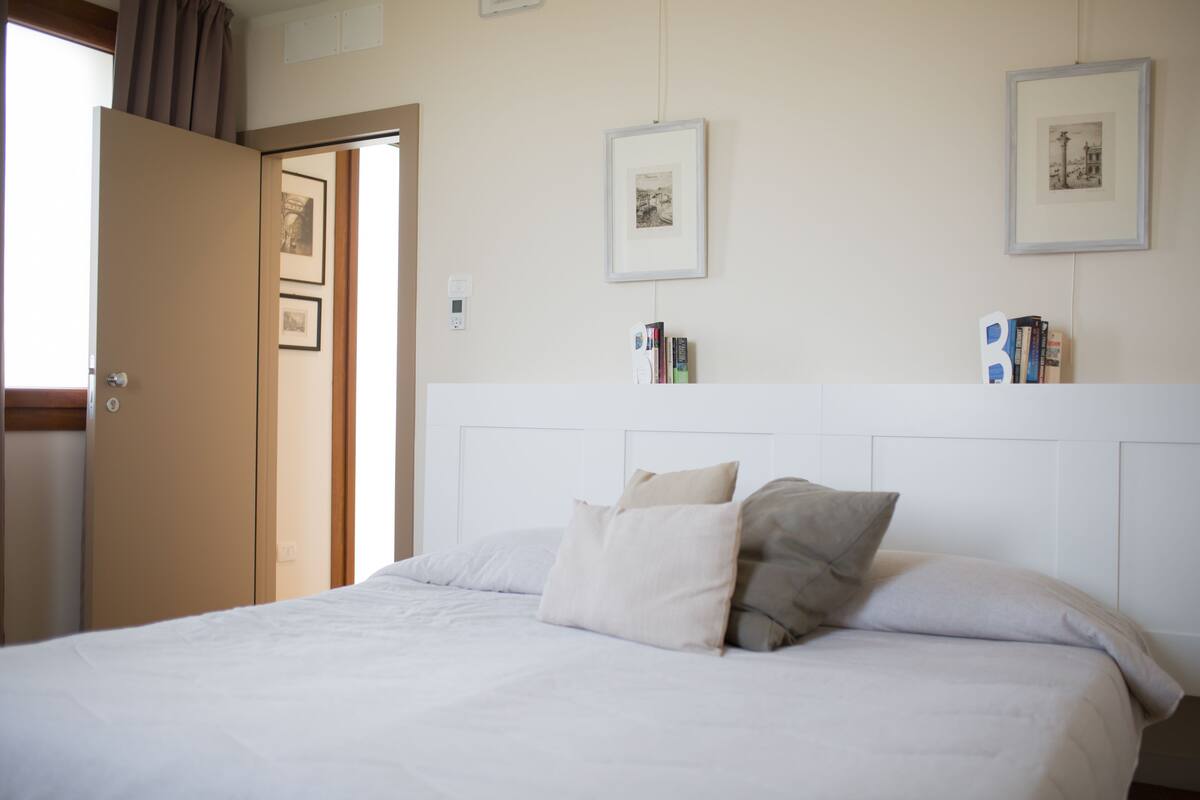A neatly made bed with pillows, a bedside table with books, and framed pictures on the wall.