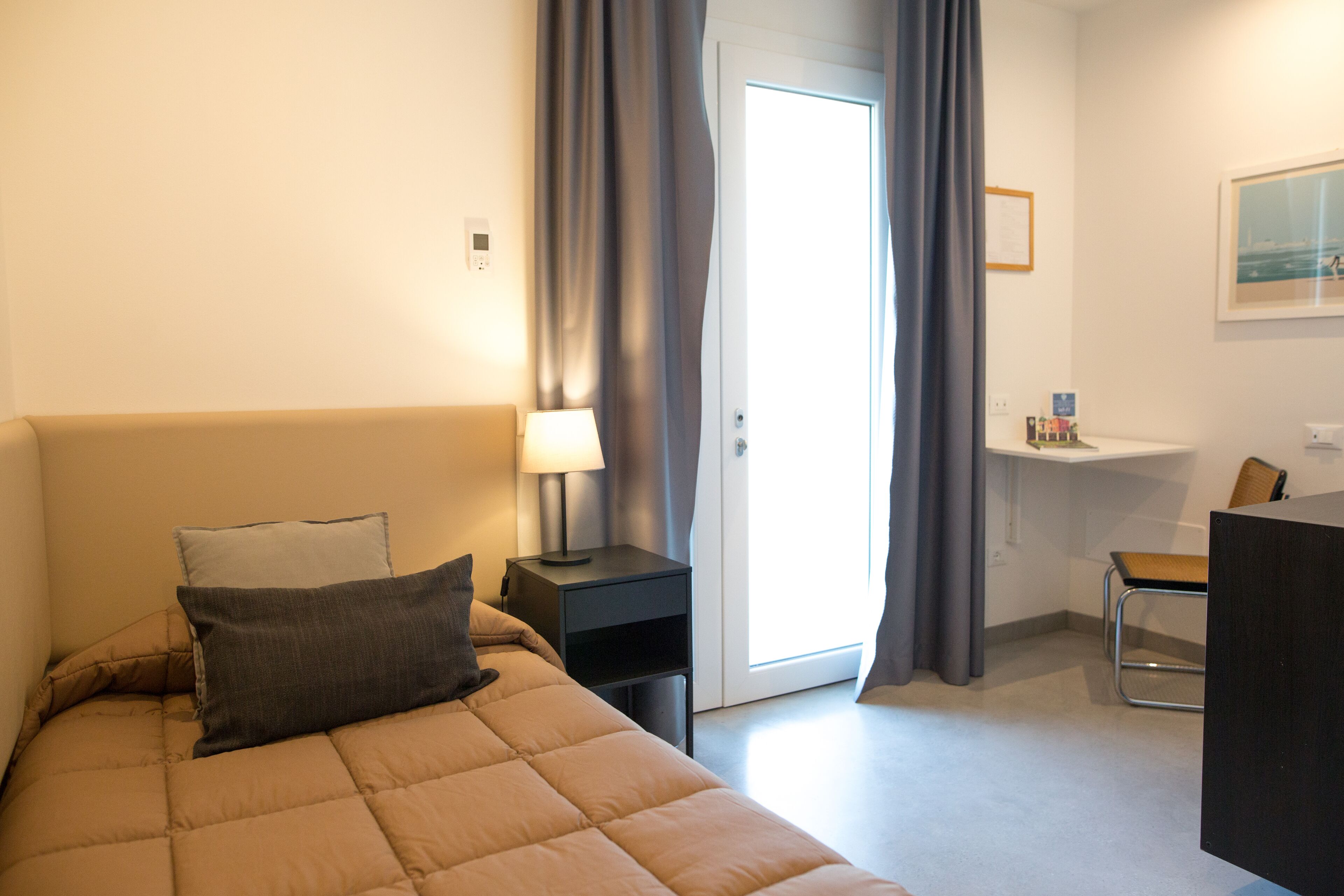 Une chambre à coucher comprenant un lit, une table de chevet, une lampe, une chaise, un bureau et une porte donnant sur un balcon.