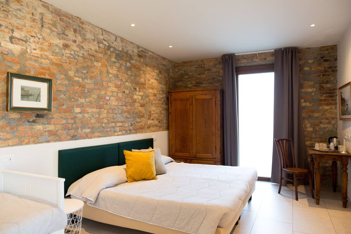 A bedroom with a brick wall, a wooden wardrobe, a bed with white bedding, a chair, and a table with a kettle and cups.
