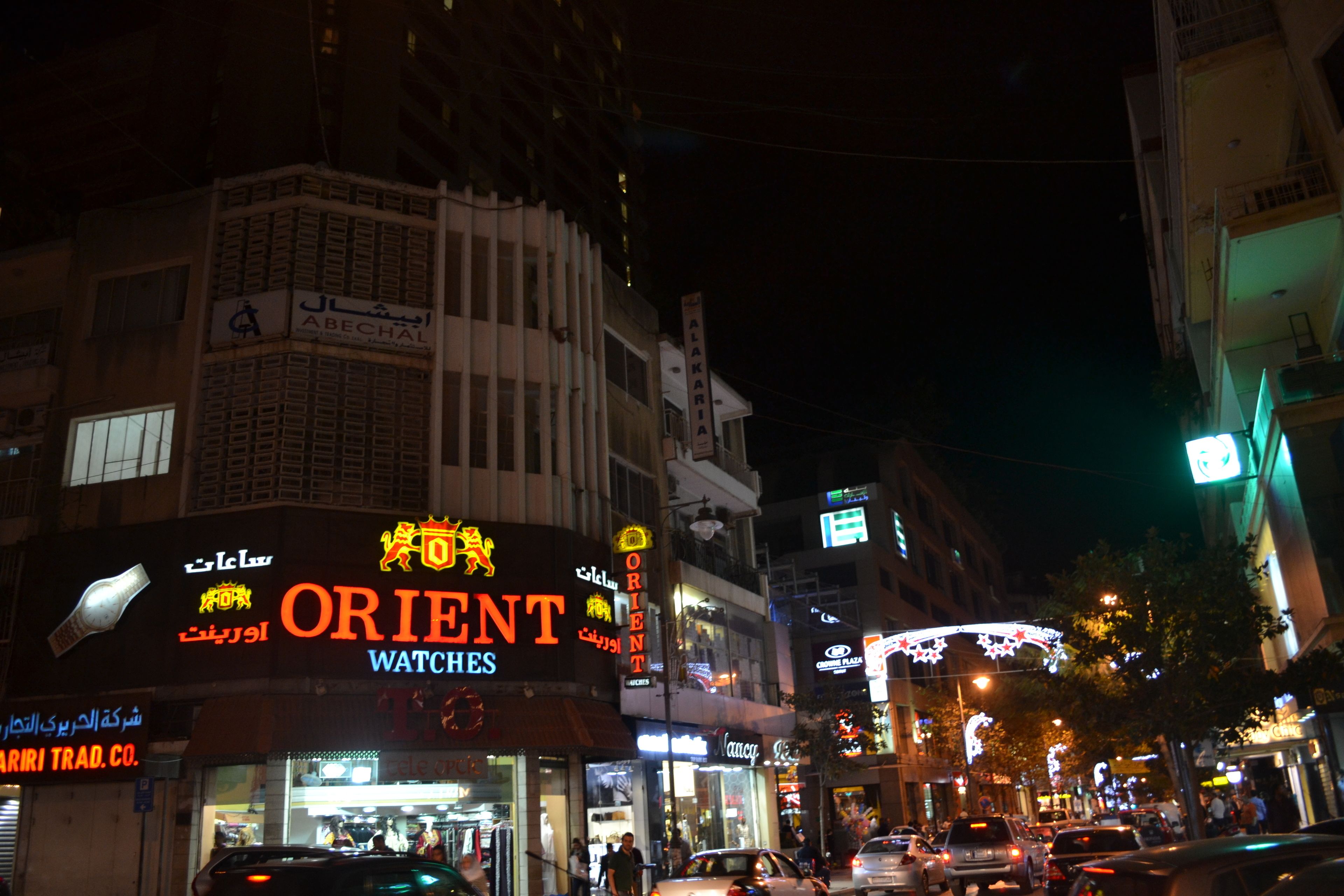 Calle iluminada de noche con escaparates, incluyendo un llamativo letrero de 'Orient Watches'.