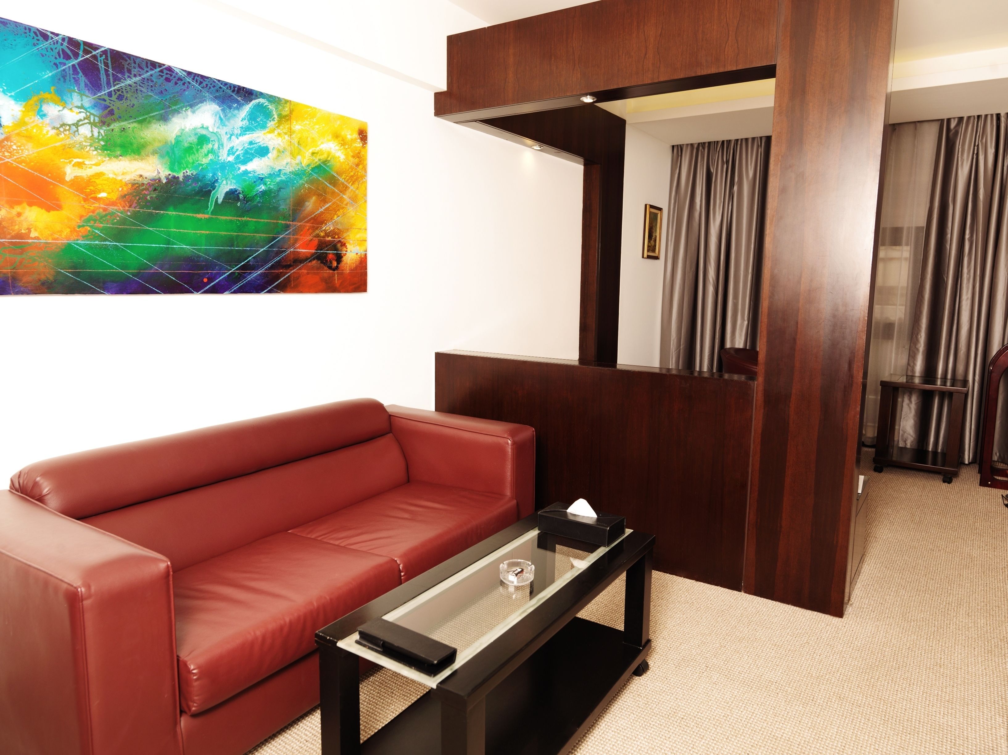 A modern living room with a red leather sofa, a glass-top coffee table, and a colorful abstract painting on the wall.
