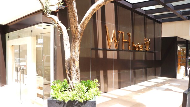 A modern hotel entrance with a glass facade, a tree planter with greenery, and a sign with the hotel's name. - WH Hotel (Beirut)