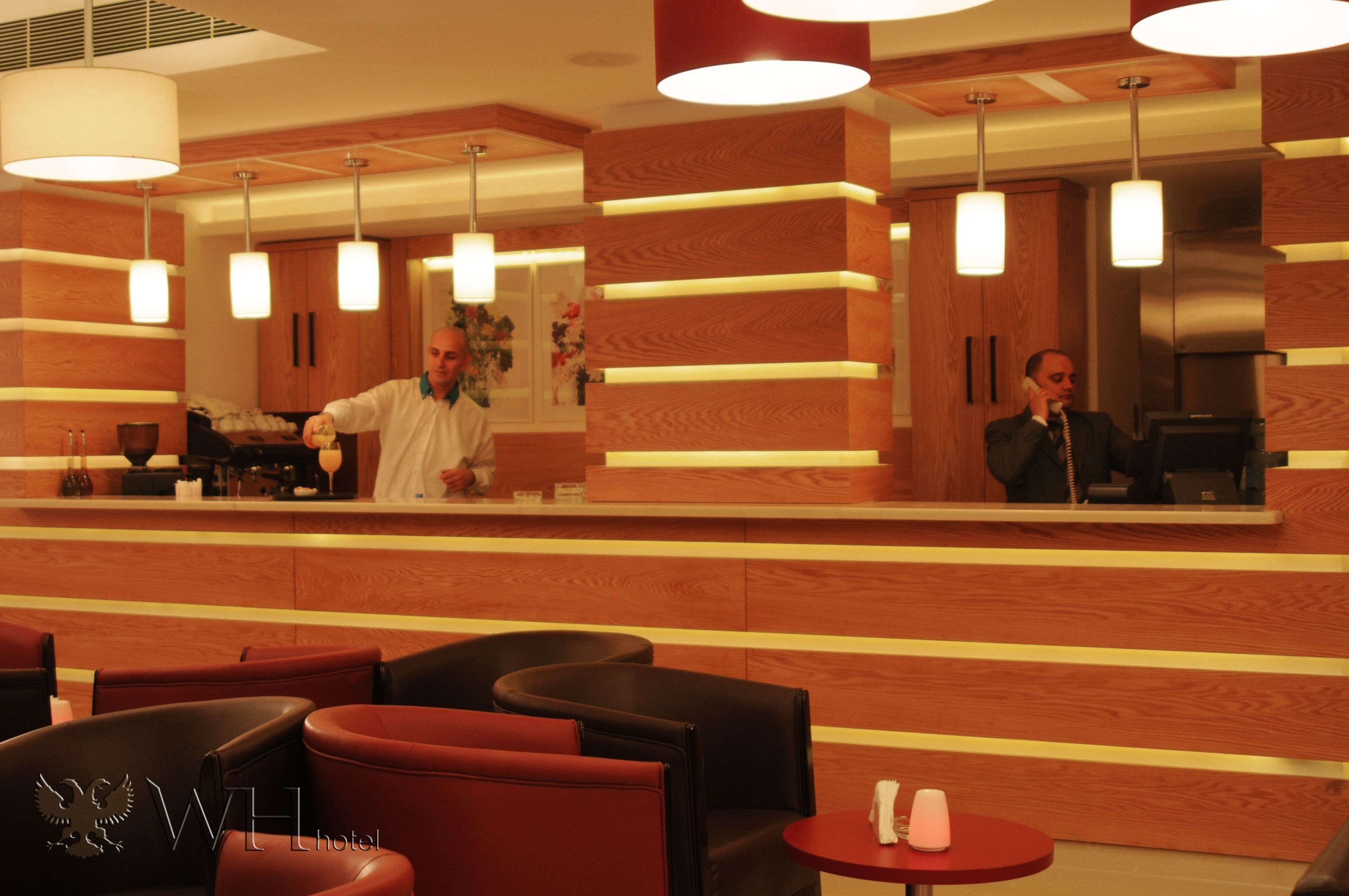A hotel lobby with a reception desk, wooden paneling, and modern pendant lighting. There are leather seating areas and a small table with a lamp.