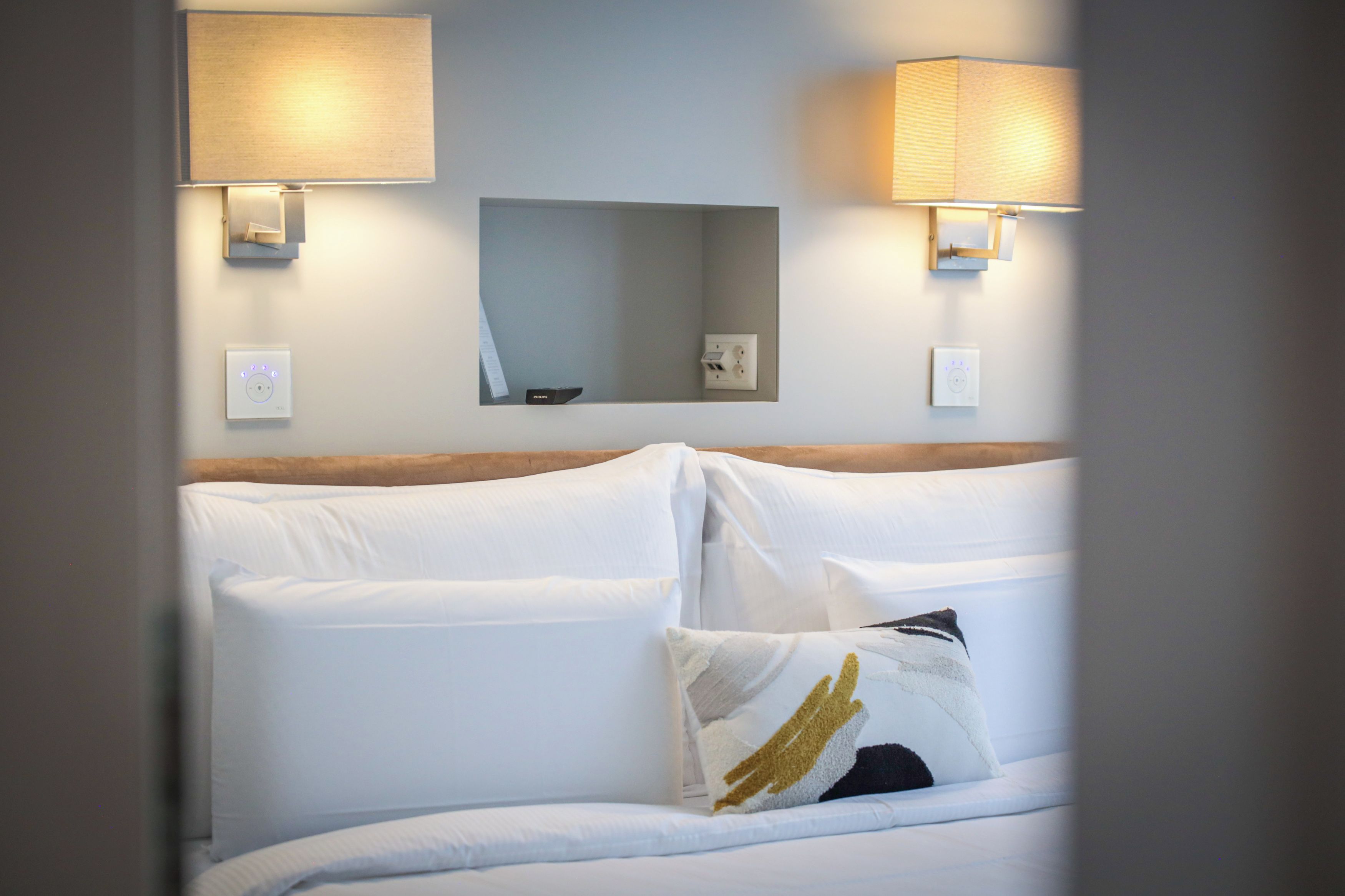 Habitación de hotel con cama, dos lámparas de pared, un enchufe y un espejo sobre la cama.