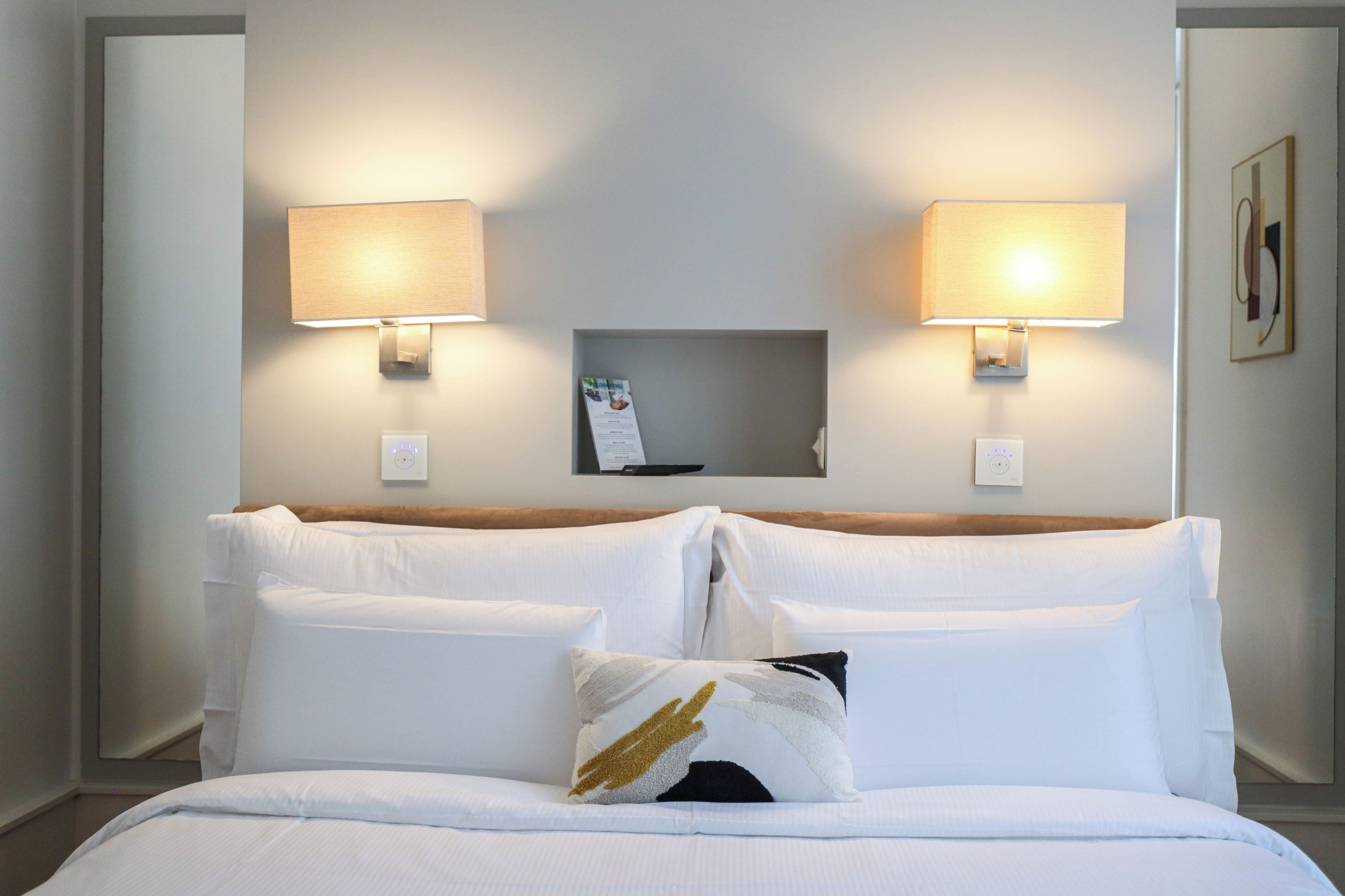 Habitació d'hotel amb un llit, dues làmpades penjades a la paret, una petita estant amb un menú i un termòstat fixat a la paret.