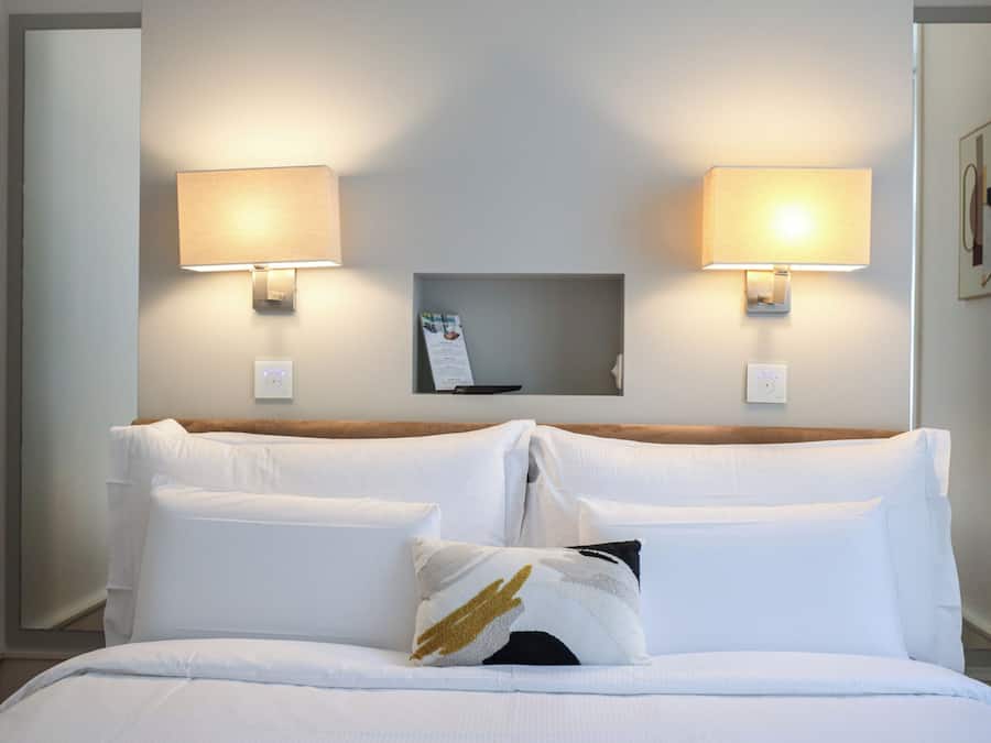 Habitació d'hotel amb un llit, dues làmpades penjades a la paret, una petita estant amb un menú i un termòstat fixat a la paret.