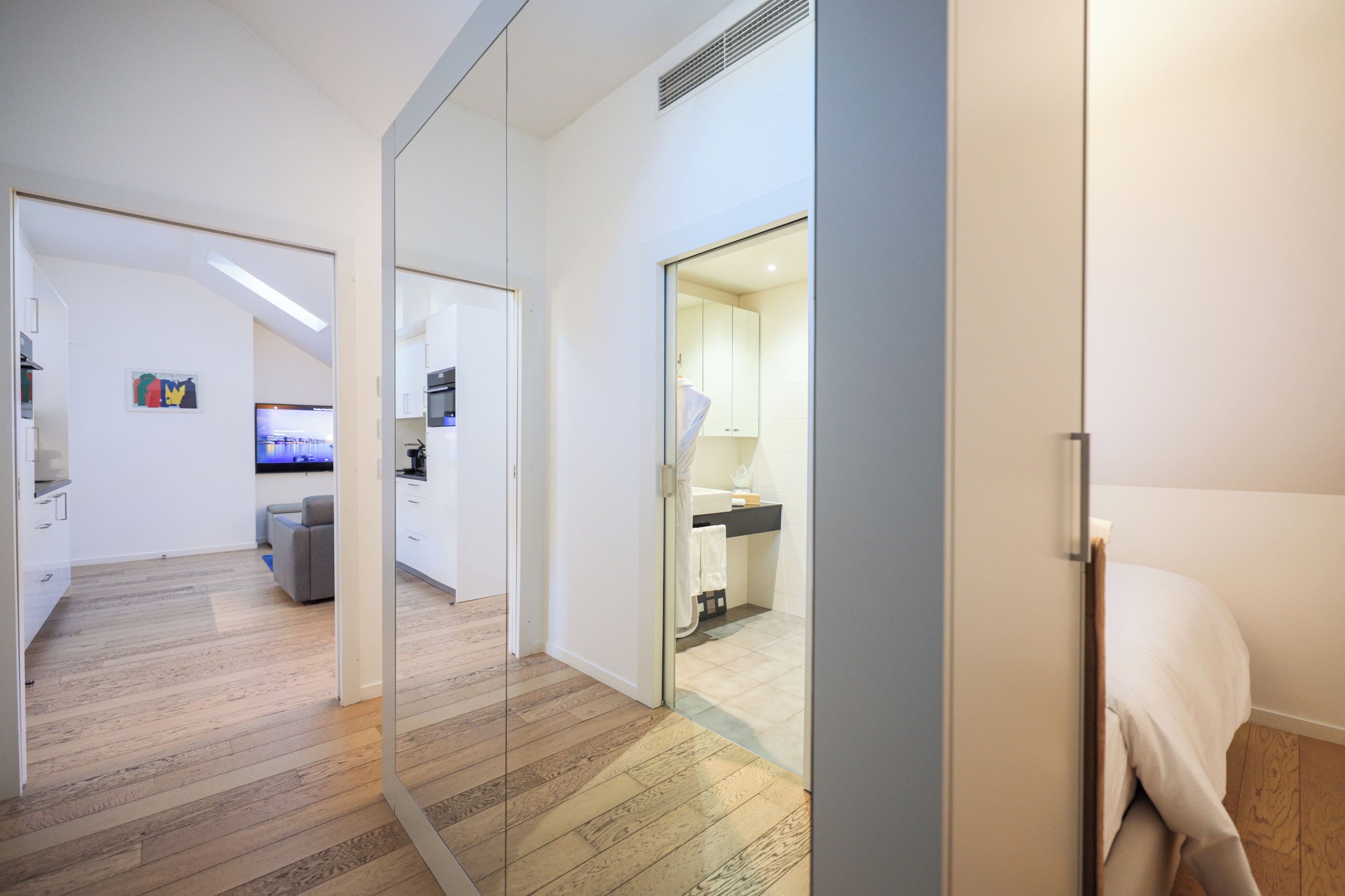 Un interior moderno con piso de madera, un sofá gris, una zona de cocina y un baño visible a través de una puerta de vidrio.
