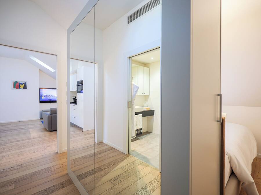 Un intérieur moderne avec un plancher en bois, un canapé gris, un coin cuisine et une salle de bain visible à travers une porte en verre.