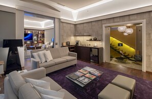 A modern living room with a sofa, coffee table, and a TV. There is a staircase with a chandelier and a bookshelf in the background.