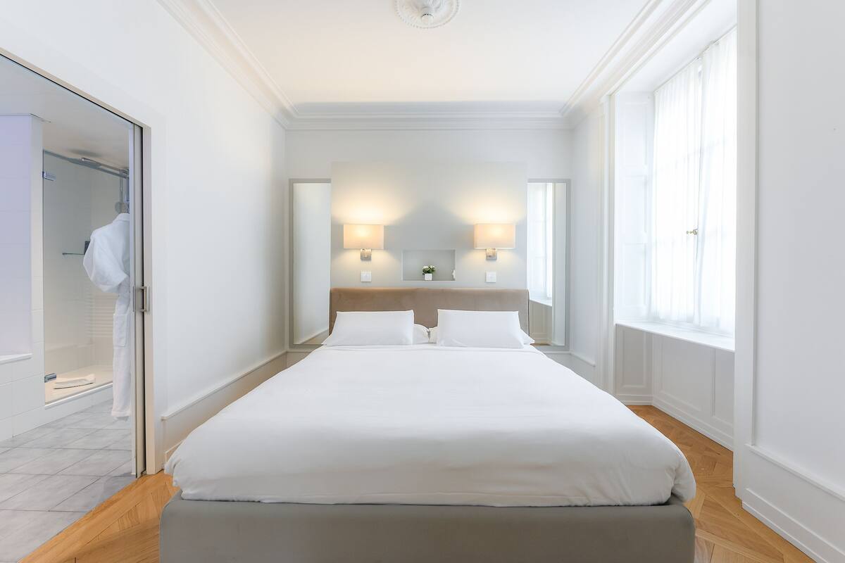 A hotel room with a bed, bedside lamps, a mirror, and a window.