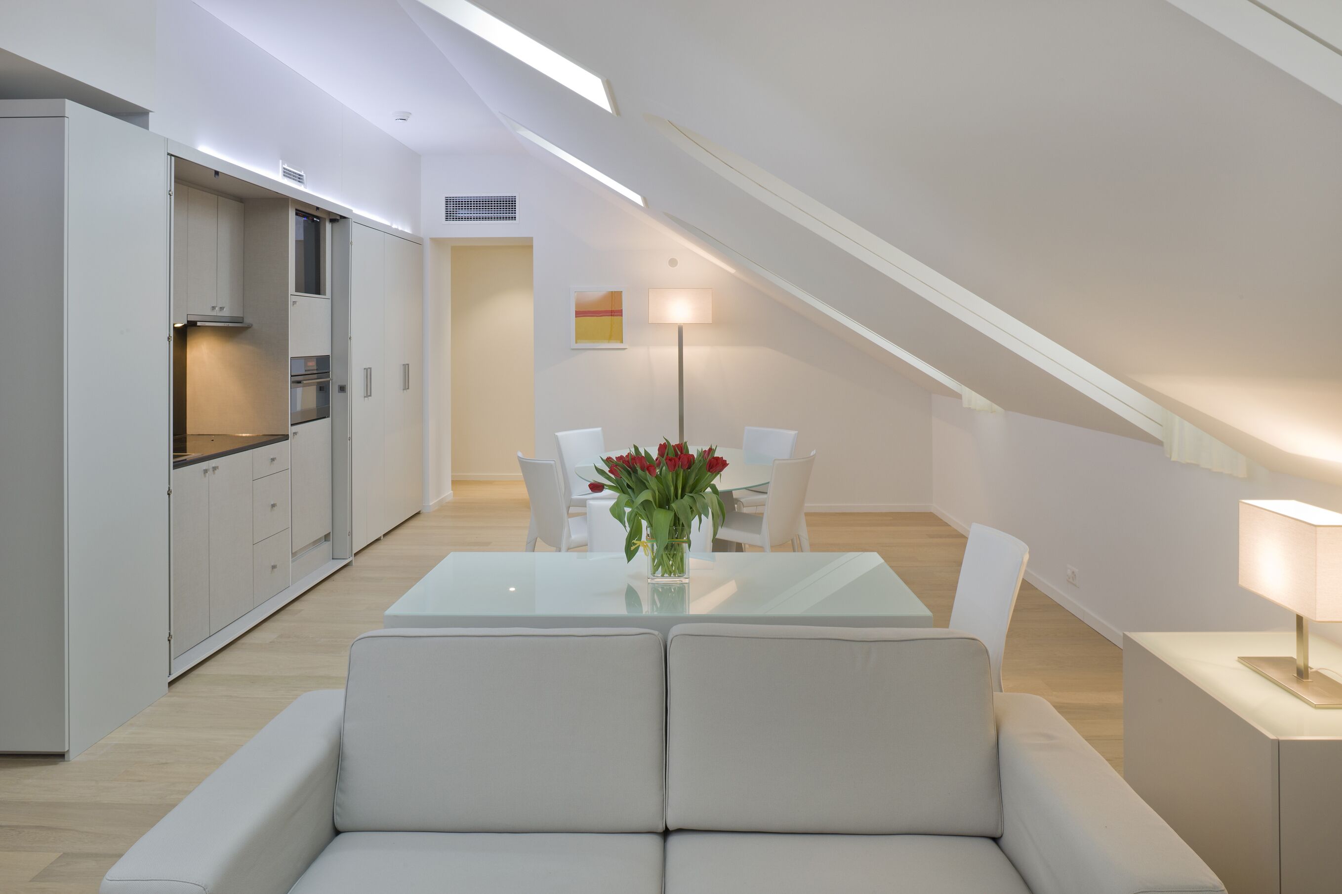 Una cocina moderna con armarios blancos, un comedor con mesa de vidrio y un rincón de estar acogedor.