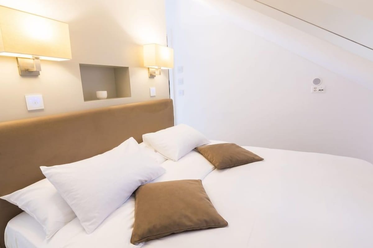 A hotel room with a bed, pillows, wall-mounted lights, and a small shelf.