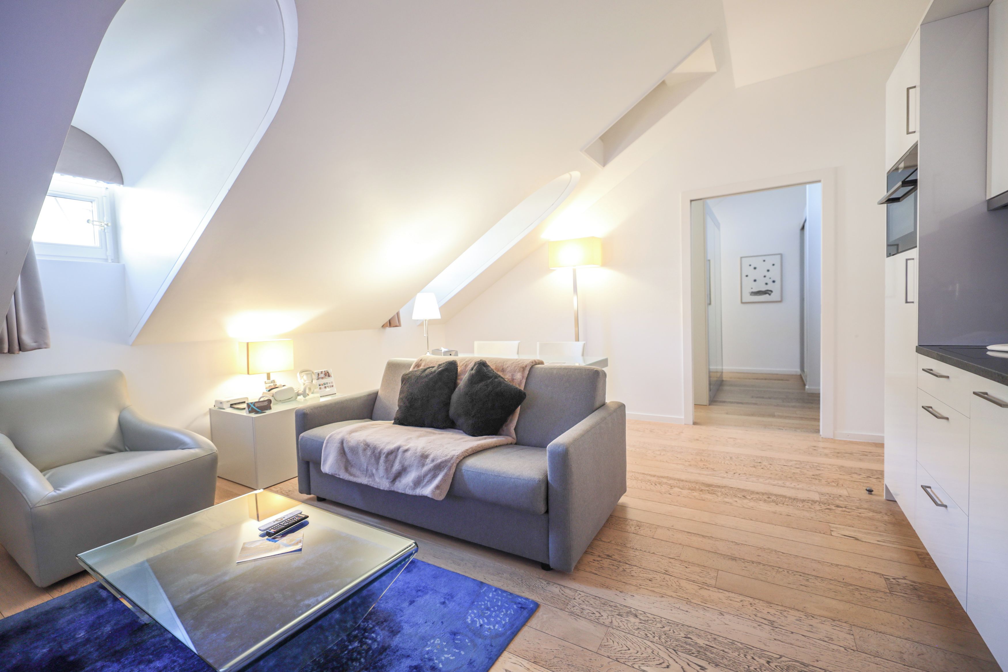 Un appartement moderne de style « loft », avec un plafond en pente, un canapé gris, une table basse et un coin cuisine.